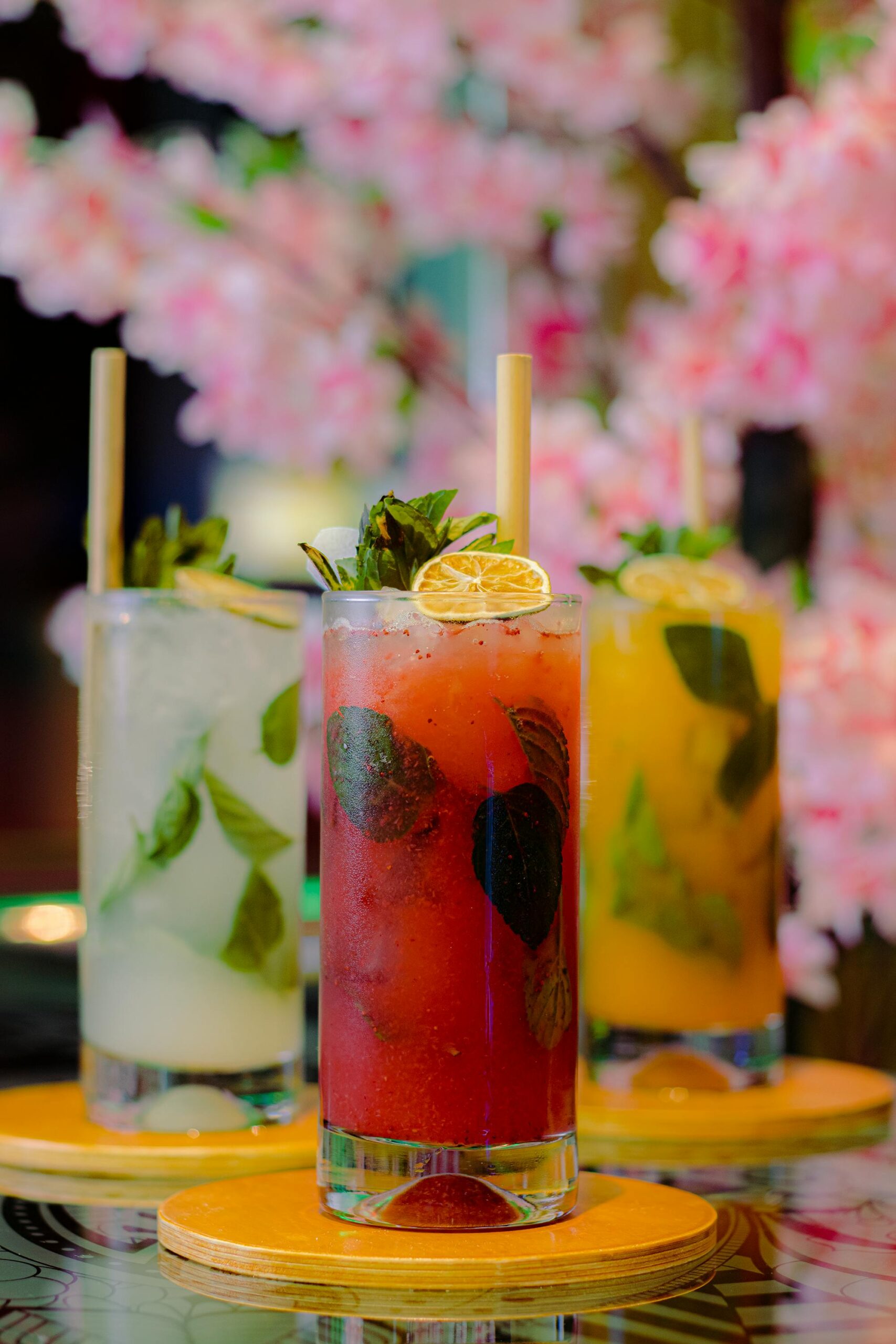 Three vibrant cocktails on wooden coasters against a backdrop of cherry blossoms, perfect for a refreshing experience.